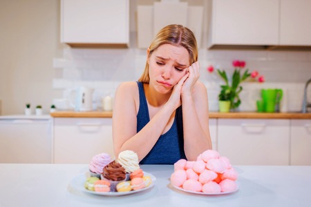 Diet struggle. Young sad woman in blue T-shirt chooses between fresh fruit vegetables or sweets while looking at them in the kitchen. Healthy and unhealthy food. Dietingの写真素材