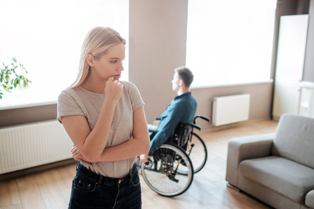 Young man sit with special needs sit on wheelchair and look on window. Woman stand in front and think. Argue and quirrel. Couple upset and unhappy.の写真素材