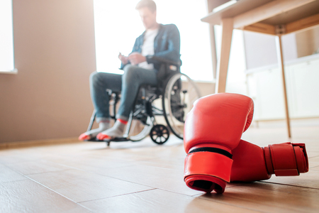 Upset young ex-sportsman with disability and inclusiveness sitting on wheelchair. Boxers gloves lying on floor. Cant use them anymore. Person with special needs.の写真素材