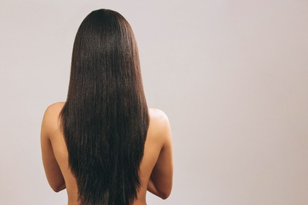 Young woman with long black hair posing on camera. Showing lenght of it. Smooth treasure. Standing back to camera. Naked. Isolated on light background.の写真素材