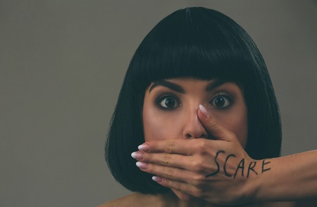 Young scared woman with black hair posing on camera. Scared afraid model look straight. Mouth closed with hand. Fear in eyes. Bob haircut. Isolated on dark background.の写真素材