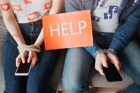 KYIV, UKRAINE - APRIL 26, 2019: Facebook logo.  illustration. Facebook icon. Young man and woman have social media addiction. Cut view of people sitting on sofa. Phone in hands. Help paper.のeditorial素材
