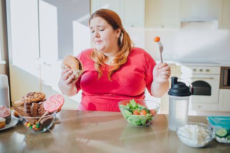 Fat young woman in kitchen sitting and eating junk food. Holding burger and tomato on fork. Regret and unhappiness. Body positive. Sweets on left. Healthy food on right side.の写真素材