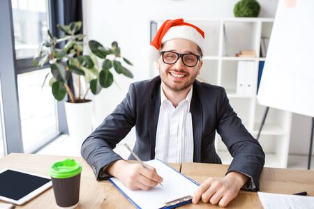 Young cheerful businessman in office. Celebrating new year or Christmas. Working alone. Writing notes. Festive mood. Positive cheerful nice man.の写真素材