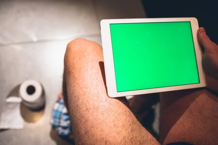 Up side view of man with hairy legs sitting on pot in rest room and hold tablet with green screen. Play and have fun alone in toilet. Paper roll on floor. Defecating.の写真素材