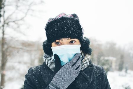 Young woman wear face mask for sickness coronavirus protection. Female person hold hand on it and think. Fear of sickness and infectioned body. Stand alone outside in warm clothes. Winter time.の写真素材