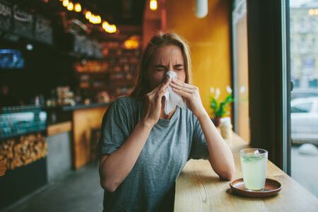 Young blonde in a cafe blows her nose. She has a runny nose. On the table is a cup of tea.の写真素材