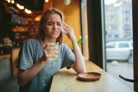 Young blonde in a cafe. She has a cold and a runny nose, her head hurts. She holds a cup of hot teaの写真素材