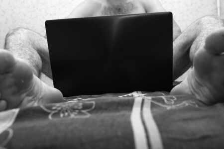 Young naked man with hairy body sit and hold laptop between legs. Cover himself. Low view. Black and white picture. Intimacy. Cut view.の写真素材