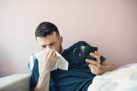 Young ill man with hanky. The guy lies in bed with a blanket sneezes and covers his nose with a napkin. In the other hand holds a cup. Isolated over light backgroundの写真素材