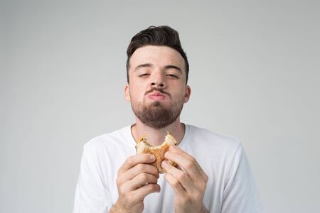Young man isolated over background. Happy satisfied guy look on camera and chewing burger. Eating bad fatning food with pleasure. Fastfood in hands.の写真素材