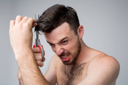 Young man isolated over background. Guy trying to cut hair with scissors by himself. Hard to do it alone. Emotional man with facial expression. Portrait of guy in action.の写真素材