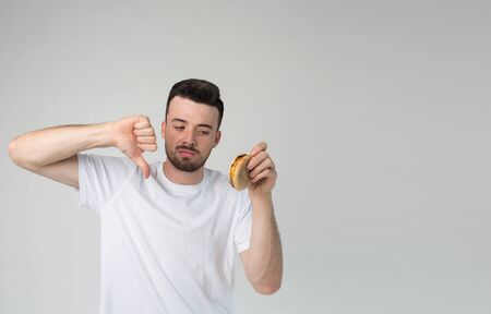 Young man isolated over background. Guy hold big thumb down for untasty burger in his hand....