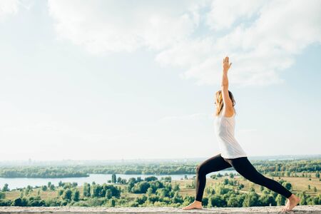 Young woman practices yoga outside. Blonde girl standing in Warrior I posture Virabhadrasana 1 , full length on parapet on beautiful view. Trees river and sky on backgroundの写真素材