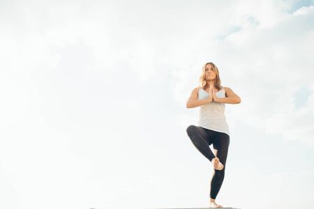 Young woman practices yoga outside. Blonde girl standing on one leg, another raised bent at knee. Hands at chest level, palms touching. Sky and clouds on background.の写真素材