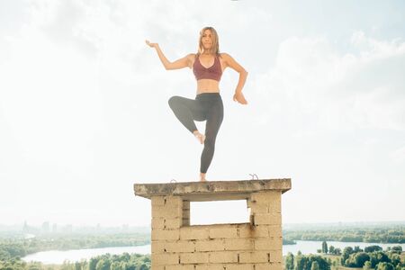 Young woman practices yoga outside. Calm girl on roof. She stand on one leg another leg bent at knee hands bent at elbows one turned up other down. Trees river and sky on background.の写真素材