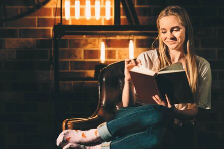 Girl in a cozy dark room reading a book with glasses. Blonde and warm lamp lightの写真素材