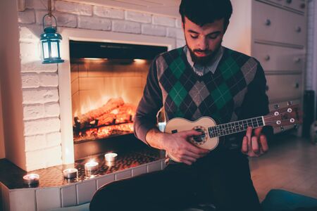 Picture of young man sitting at fireplace and playing ukulele. Alone in room inside practicing. Singing songs and playing melodies.の写真素材