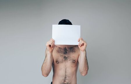 Young man isolated over background. Naked ordinary guy cover face with white blank page. Hide form somebody. Posing on cameraの写真素材