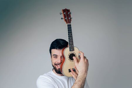 Young man isolated over background. Portrait of guy cover face part with small ukulele. Musician and player have fun alone during posingの写真素材