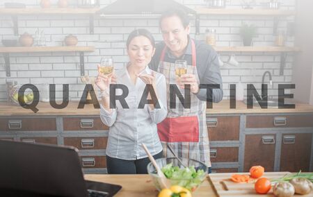 Cheerful couple stand together in kitchen. They hold glasses of wine and look on laptop. People smile. They have vegetables on table.の写真素材