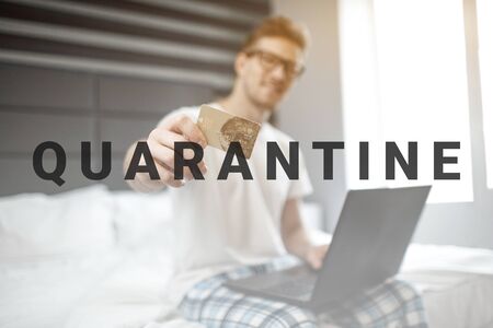 Nice young man sit on bed early morning. He show credit card on camera. Guy hold laptop. Electronic moneyの写真素材