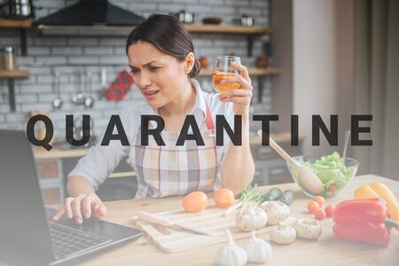Disturbed woman sit on table in kitchen. She hold glass of wine and type on keyboard. Model look on laptop screen. She is emotional.の写真素材