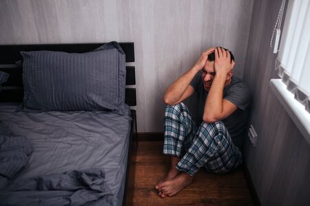 Depression, panic attack concept. A man is sitting in a corner near the bed. He feels bad.の写真素材