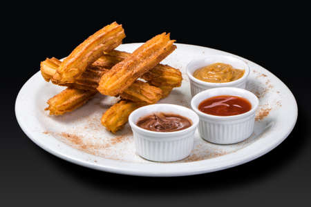 Churros with sugar and cinnamon served with chocolate, milk caramel and guava jam served in a white dish on black background.の写真素材