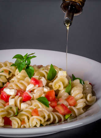 olive oil poured into pasta salad with chopped tomatoes and basil on light gray background.の写真素材