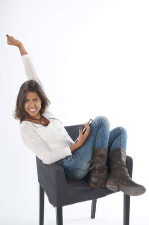 Happy young girl with her arm up, wearing casual clothing, on white, sitting on sofa holding tablet PC with hands up.の写真素材