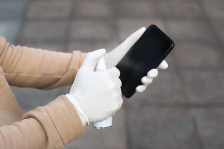Young woman disinfects her phone with a hand sanitizer. Woman holding a mobile phone in her hands wearing latex protective gloves at the street in public place.の写真素材