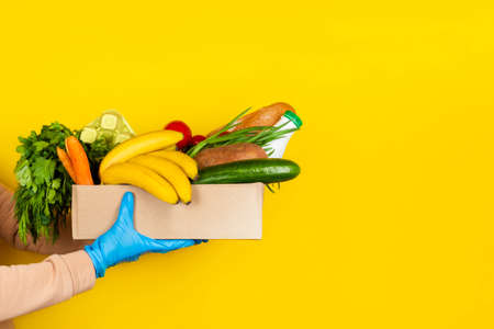 Safe food delivery, online shopping or donation concept. Food delivery during virus quarantine. Box with different food ingredients in female hands in disposable glovesの写真素材
