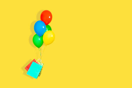 Paper bags and a bunch of balloons fly. Sale, present, delivery concept. Green, red, yellow, and blue Balloons, red and blue paper bags flying isolated on yellow background. Banner with copy space.の写真素材
