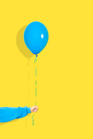 Female hand hold blue balloon. Party or present concept. Blue Balloon and hand in blue hoodie isolated on yellow backgroun.の写真素材