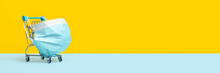 Shopping cart or trolley with protective medical mask against coronavirus. Safe and online shopping on quarantine concept. Bright yellow background, long banner with copy space.の写真素材