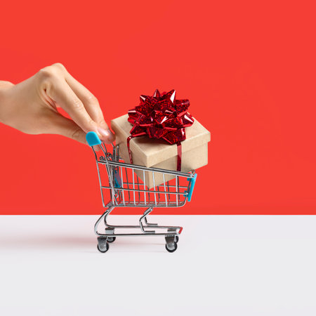 Hand pushing Toy shopping cart or trolley with large gift box. A gift for a holiday, anniversary, or birthday. Trolley cart with a gift box Isolated over red backgroundの写真素材