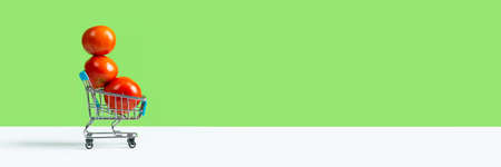 Toy shopping cart or trolley full of large red tomatoes isolated on green background. Long Fall harvest sale concept. Banner with copy space.の写真素材