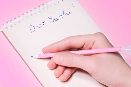 Dear Santa letter concept. Hand holding pink pen decorated with feather and writing in Notepad...