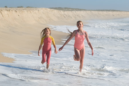 Two girls are running joyfully along the shoreline, splashing through the shallow waves on a sunny beach. The sandy dunes and clear sky create a perfect summer day atmosphere full of fun.の写真素材