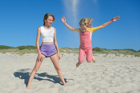 Two girls are playing on a sandy beach. One is jumping joyfully as sand flies up, while the other stands nearby, smiling. The clear blue sky and greenery create a peaceful summer backdropの写真素材