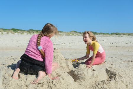 Two girls building a sandcastle on the beach under a clear blue sky. Two happy sisters having fun on the Atlantic ocean deserted beach.の写真素材