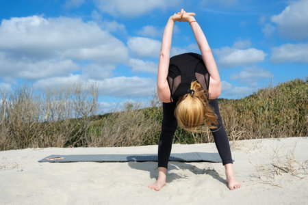 Woman practicing yoga outdoors on sandy surface, performing forward bend with clasped hands behind back, embracing balance, flexibility, and mindfulness under bright sky and natureの写真素材