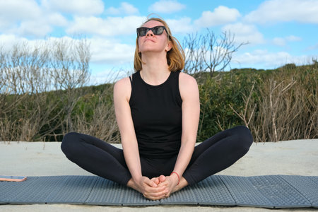 Woman practicing yoga outdoors, sitting in butterfly pose on a mat, enjoying fresh air, natural surroundings, and blue sky, embracing mindfulness and relaxation.の写真素材