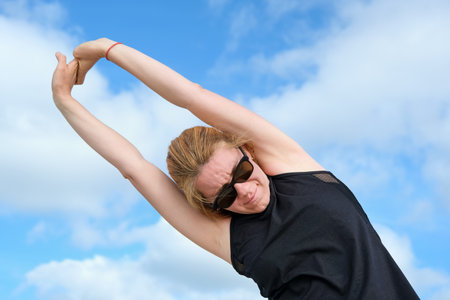 Woman stretching arms overhead outdoors under bright sky, enjoying fitness and wellness routine,...