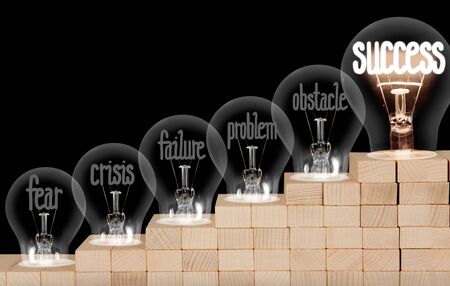 Group of shining and dimmed light bulbs on wooden block ladder with fibers in a shape of Fear, Crisis, Failure and Success concept words isolated on black background.の写真素材