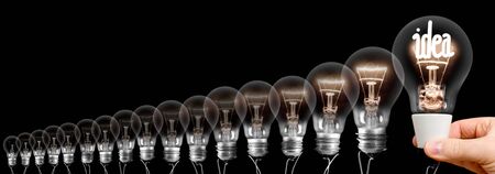 Group of light bulbs going from dark one to a shining one with fiber in Idea shape being held by a human hand isolated on black background. Concept of Thinking, Genius, Success and Creativity.の写真素材