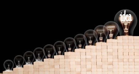 Group of light bulbs going from dark one to a shining one with fiber in Idea shape on wooden block ladder isolated on black background. Concept of Thinking, Success and Creativity.の写真素材