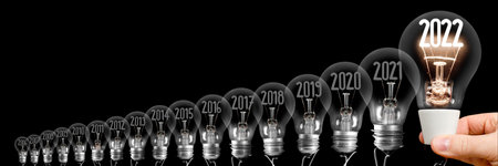 Horizontal group of shining light bulb with fiber in a shape of New Year 2022 and dark light bulbs with years passed isolated on black background.の写真素材