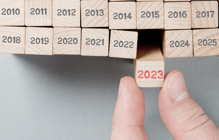 Photo of wooden blocks hold by a human hand with gray numbers and red number 2023 imprinted on...